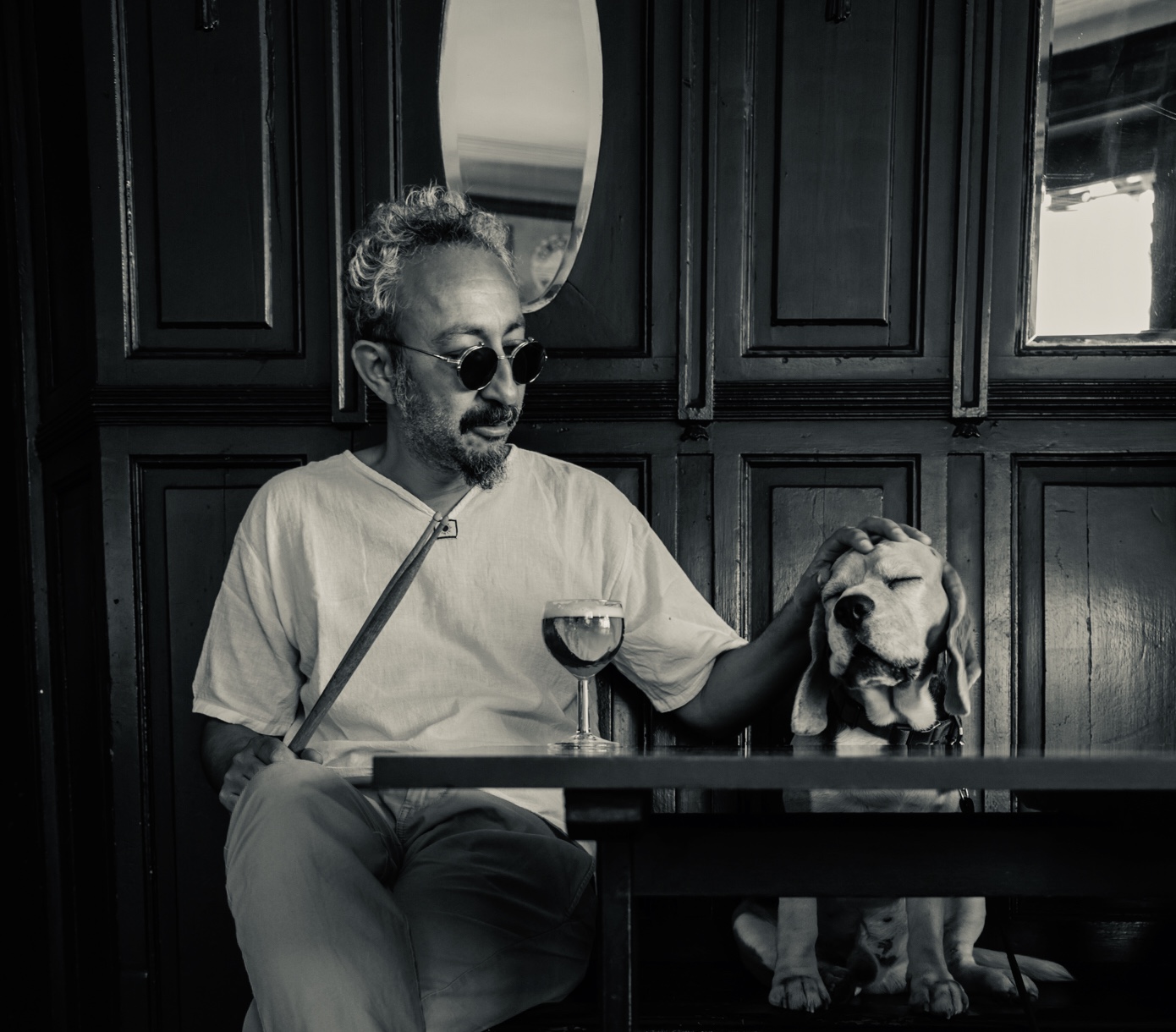 Portrait noir et blanc de MoAdib Garti assis à la table d'un bar, tenant une paire de baguette d'une main et caressant un chien de l'autre, devant un verre de vin