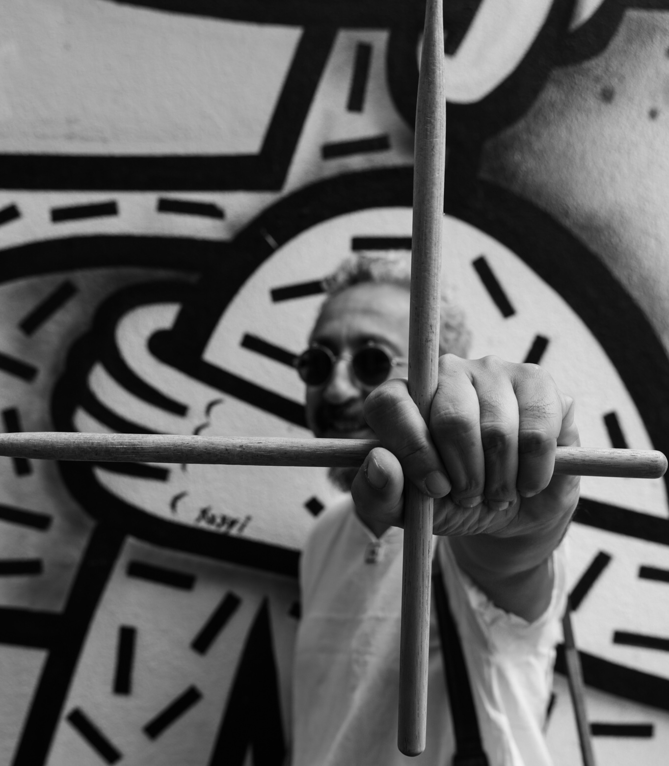 Portrait noir et blanc de MoAdib Garti posté devant un grafitti moderne et pointant une paire de baguette de batteur tenues à angle droit vers l'avant